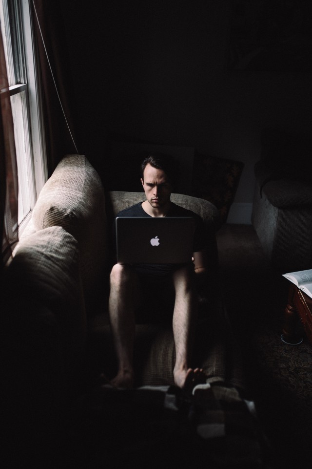 Young man is lying on the couch and working on his laptop. It's pretty dark in his room, he is barefoot and looks stressed/tense.